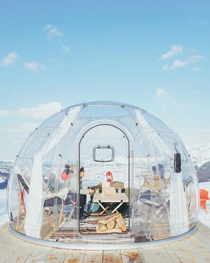 A transparent dome structure with a table and chairs inside, set on a wooden deck with a snowy landscape in the background. - SnowHouse Memorie (Minamiuonuma)