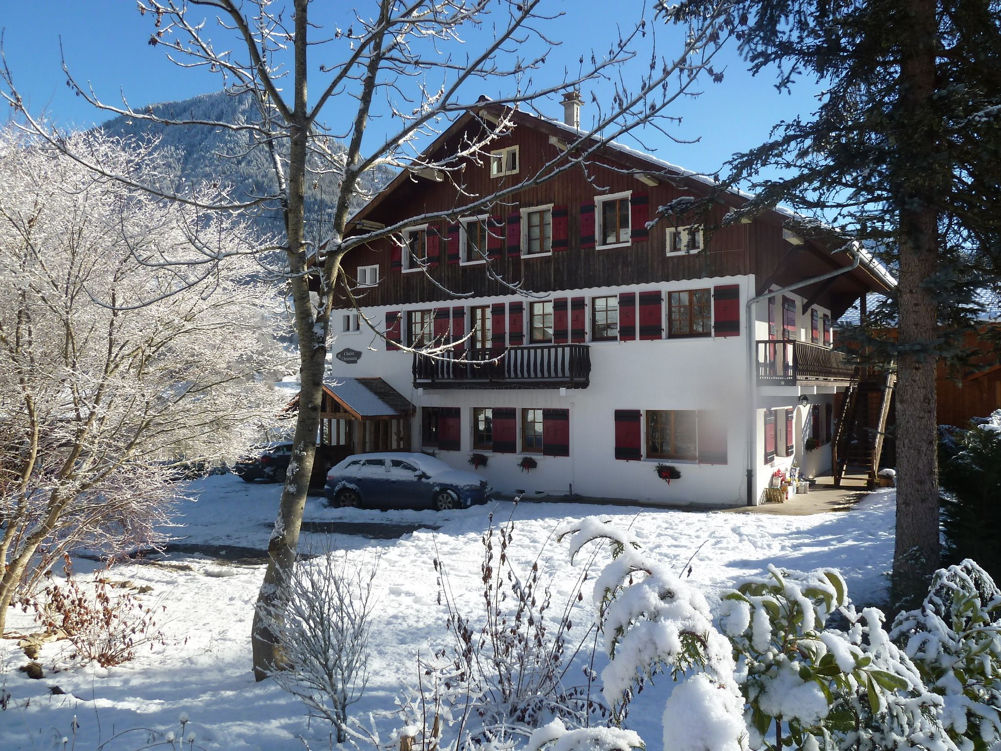 A two-story chalet with a balcony, surrounded by snow-covered trees and a clear sky.