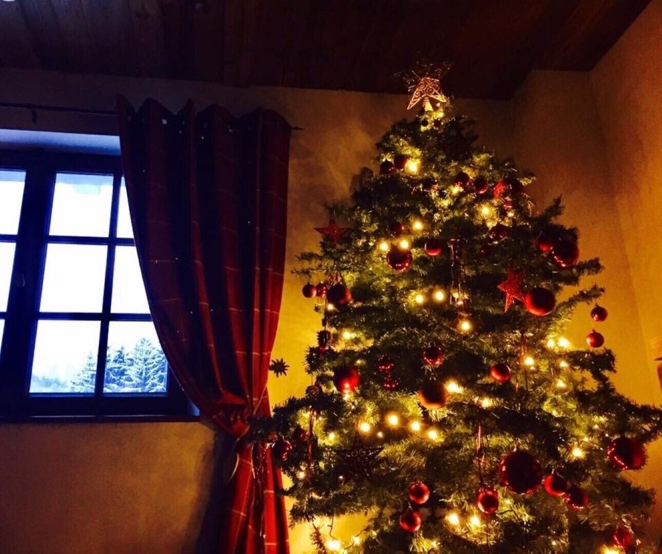 A Christmas tree with lights and ornaments, positioned next to a window with curtains.