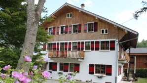 Une maison en bois à deux étages, avec des volets rouges, un balcon et un petit jardin fleuri.