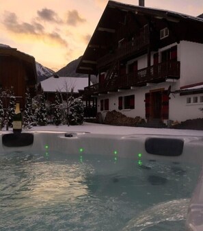 Relax in a serene winter wonderland with a hot tub in a peaceful courtyard surrounded by traditional chalet-style buildings.