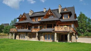 Ein großes, mehrstöckiges Holzhaus mit steinernen Fundament, mehreren Schornsteinen und einem Balkon mit aufwendiger Balustrade.