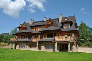 A large, multi-story wooden house with a stone foundation, multiple chimneys, and a balcony with intricate railings.