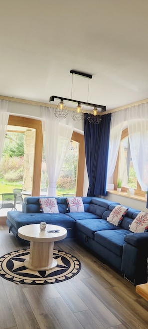 Ein modernes Wohnzimmer mit einem blauen Ecksofa, einem runden Couchtisch und einem großen Fenster mit durchsichtigen Vorhängen.