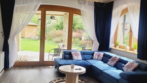 Ein modernes Wohnzimmer mit einem blauen Ecksofa, einem kleinen runden Tisch und einer großen Glasschiebetür, die zu einem Garten führt.