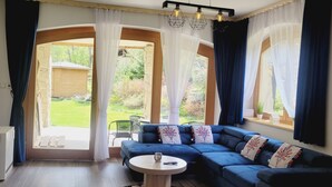 Ein modernes Wohnzimmer mit einem blauen Ecksofa, einem kleinen runden Tisch und einem an der Wand befestigten Fernseher. Große Fenster mit transparenten Vorhängen lassen viel Tageslicht herein.