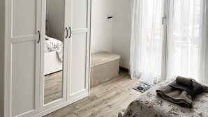 A bedroom with a large wardrobe, a bed with a floral duvet, a radiator, and sheer curtains on a window. - Sicily Bedda vacation home, apartment in the city center and a stone's throw from the sea! (Pozzallo)