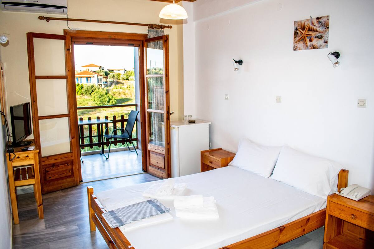 Une chambre avec un lit, un bureau avec un ordinateur, une chaise, une petite table, une table de chevet et une vue sur l’extérieur.