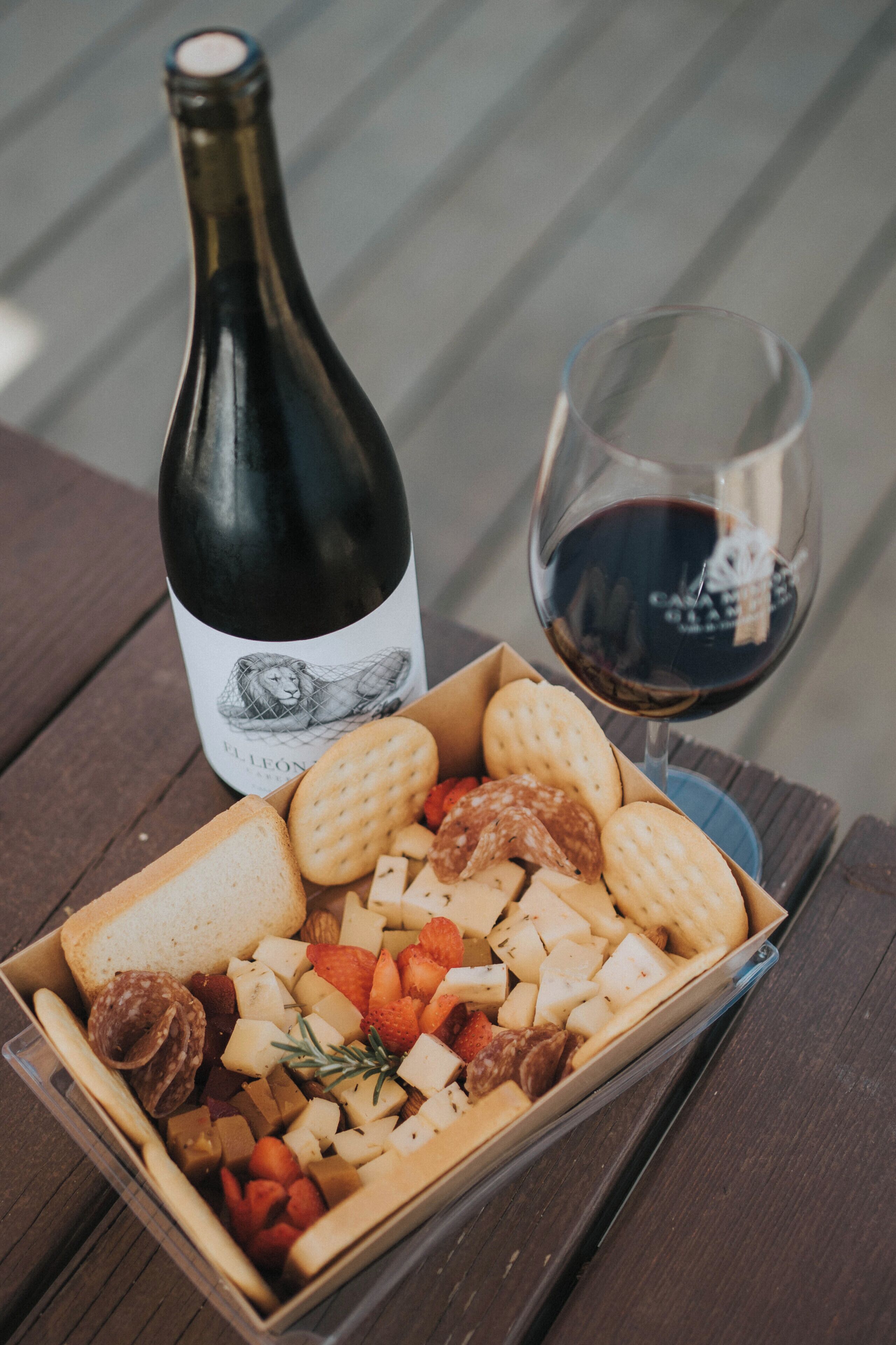 A bottle of wine, a glass of wine, and a box of assorted snacks on a wooden table.