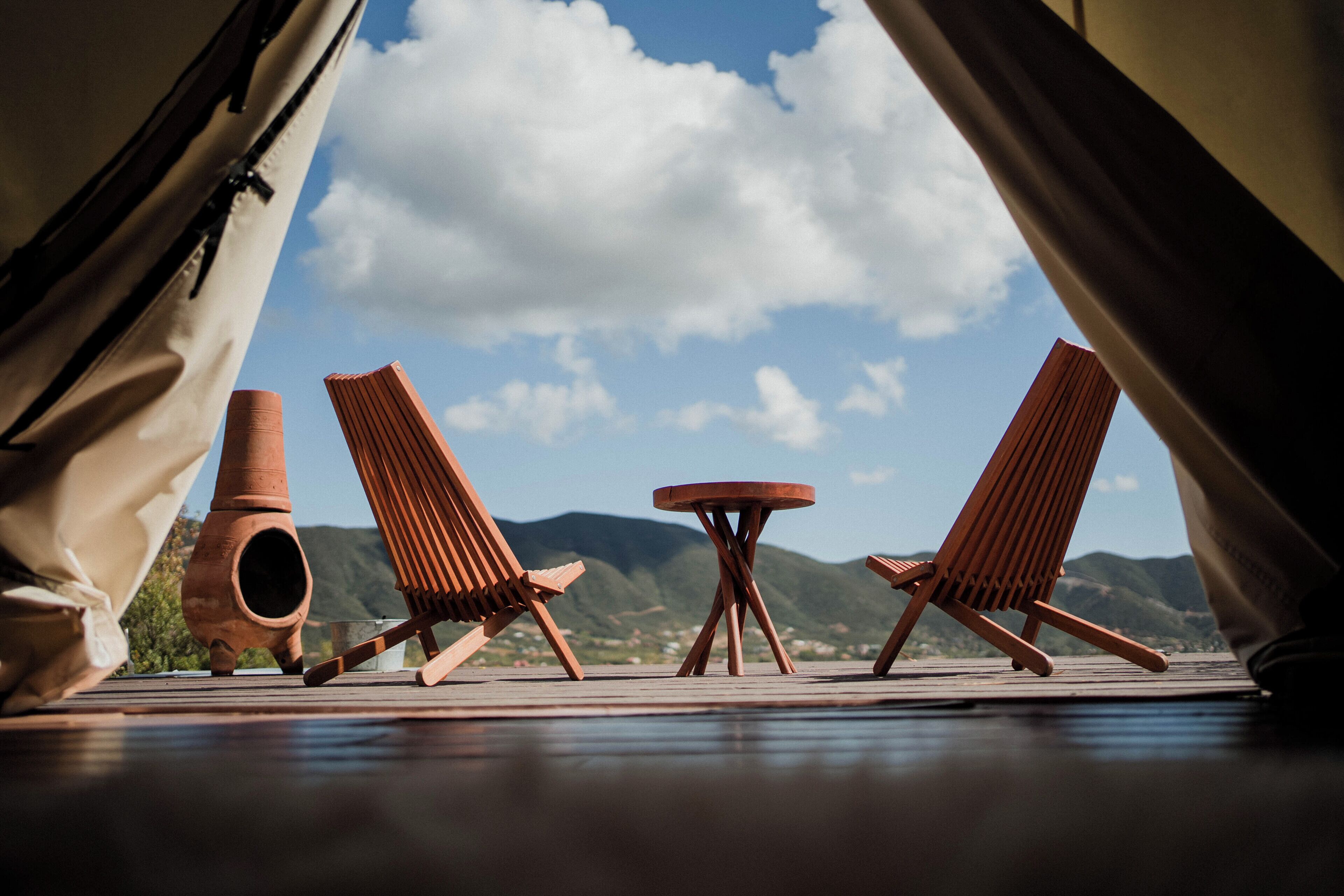 Een gebied met tenten, met twee houten stoelen, een tafeltje, een terracotta pot en uitzicht op bergen en de lucht.
