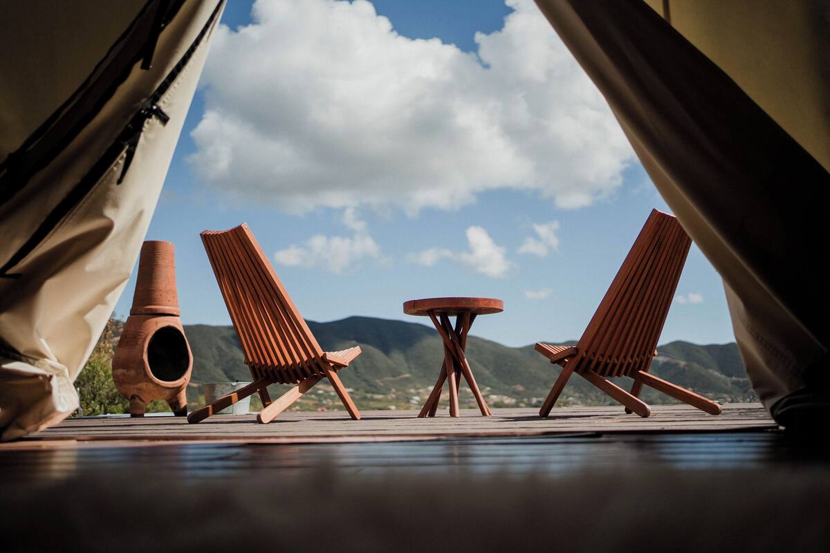 A tented area with two wooden chairs and a small table, a clay pot, and a view of mountains and sky.