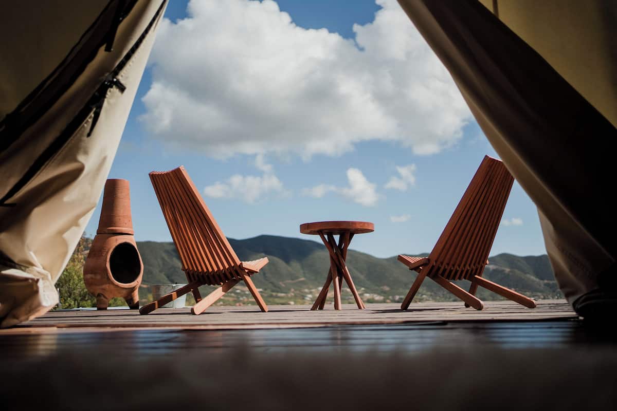 A tented area with two wooden chairs and a small table, a clay pot, and a view of mountains and sky.