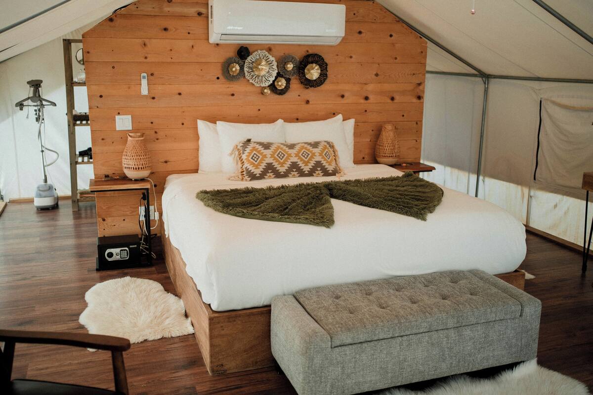 A tented room with a bed, a grey ottoman, a wooden headboard, and a wall-mounted air conditioner.