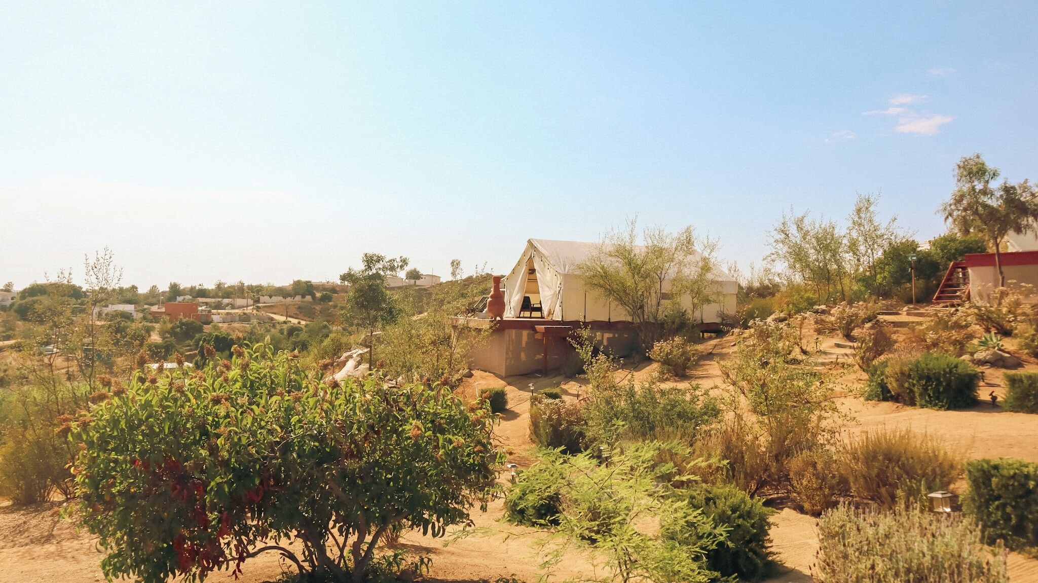A desert landscape with a tent-like structure, surrounded by sparse vegetation and a distant town.