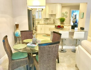 A dining area with wicker chairs and a glass table, a kitchen with white cabinets, and a bar area with white bar stools.