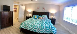 A bedroom with a bed, two bedside tables, a TV on a dresser, and a window with blinds.