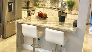 A modern kitchen with a stainless steel refrigerator, a microwave, and a marble countertop. There are two white bar stools and a decorative metal sculpture.