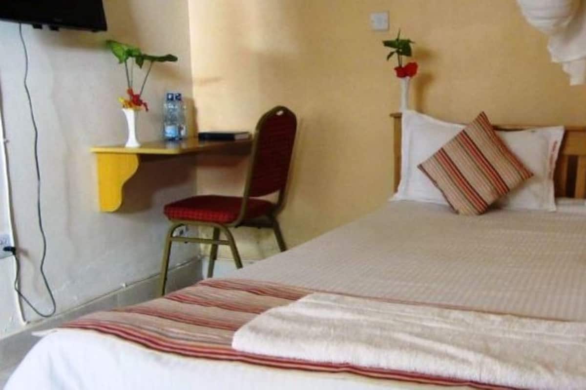 Quarto de hotel com cama, almofadas, cadeira, mesinha com um vaso e uma garrafa, e uma TV fixada na parede.
