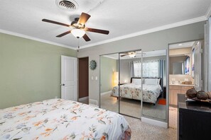 Une chambre à coucher avec un lit, un ventilateur de plafond, un miroir et une salle de bain visible à travers une porte en verre.