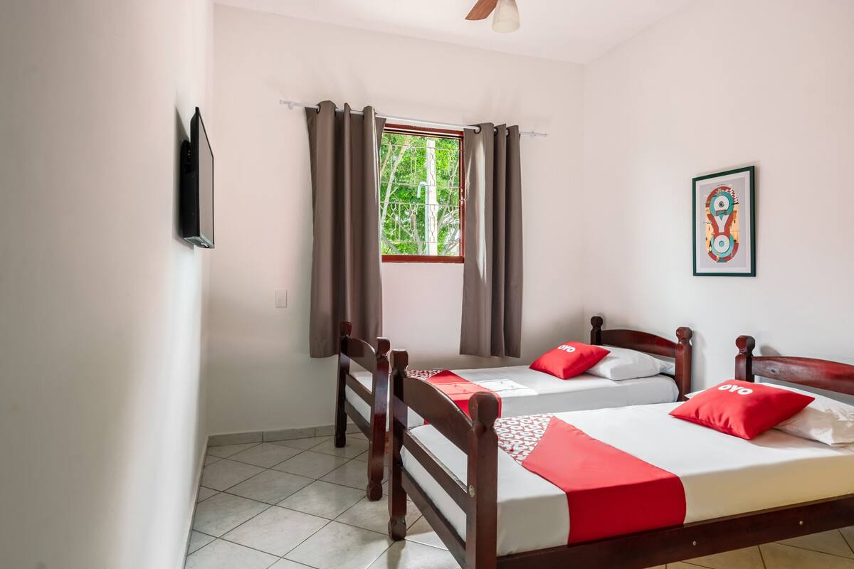 A room with two wooden beds, a window with curtains, a ceiling fan, and a framed picture on the wall.