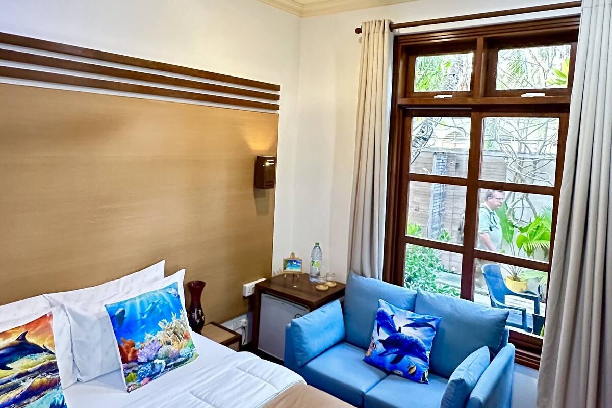 Una habitación de hotel con una cama, un sofá azul, un ventilador de techo, una ventana con cortinas y una pequeña mesa con una botella.