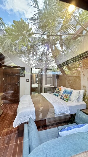 A room with a bed, a sofa, a small table, and a bathroom visible through a glass door. - Beach Cottage (Rasdhoo)