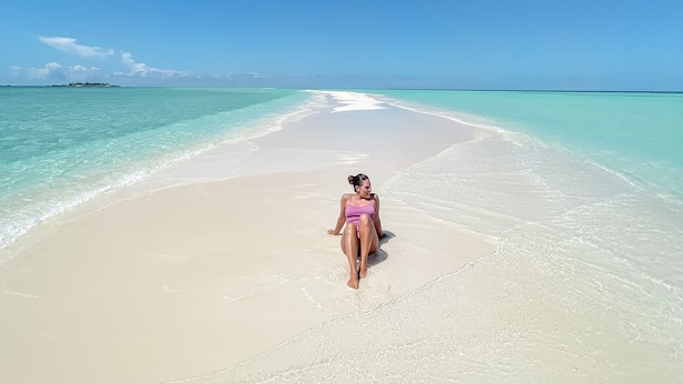 Une personne se promène sur une plage où l’eau est d’un bleu turquoise limpide et le ciel d’un bleu azur.