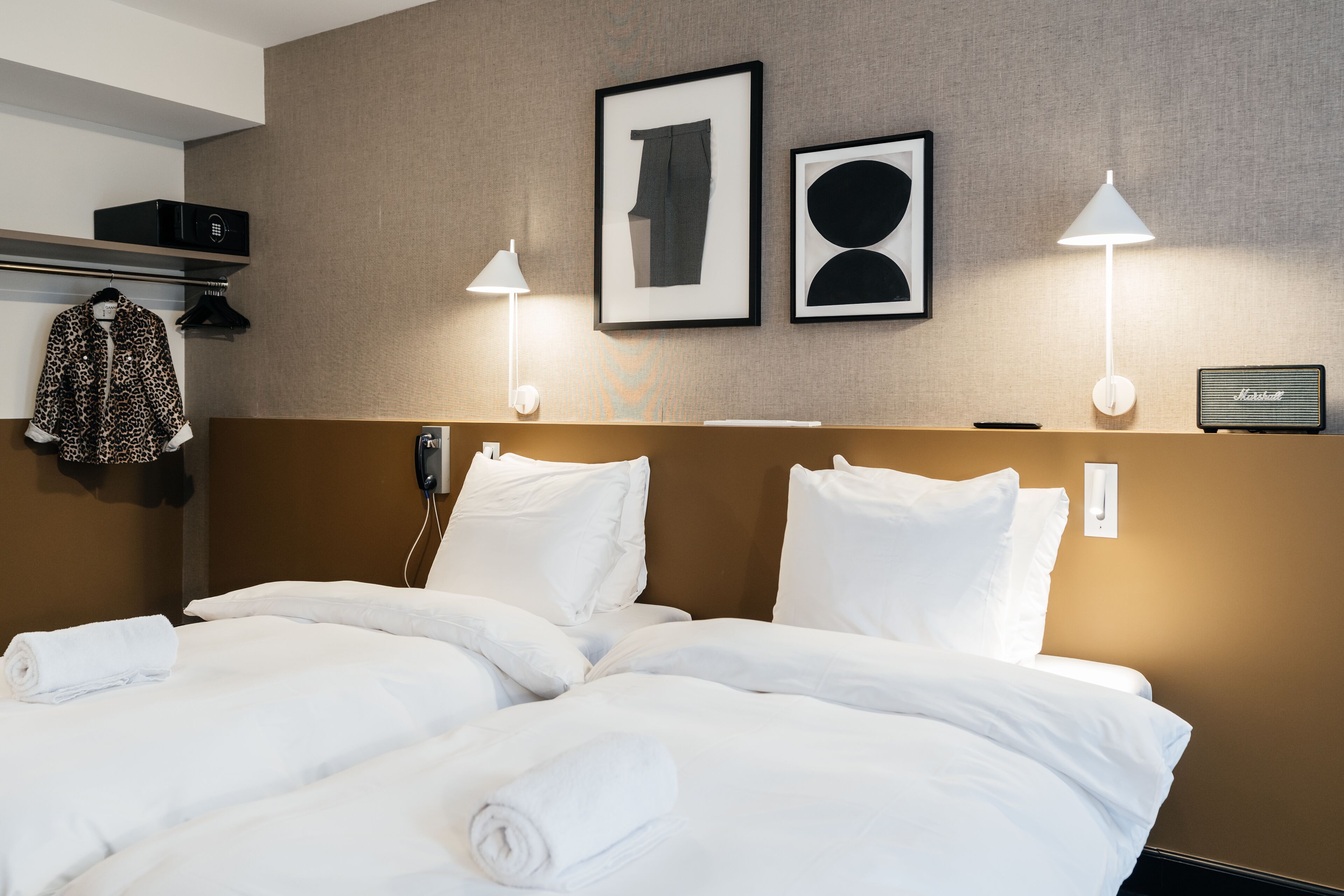 A hotel room with a neatly made bed, two wall-mounted lamps, a framed picture, a radio, and a coat rack with a jacket.