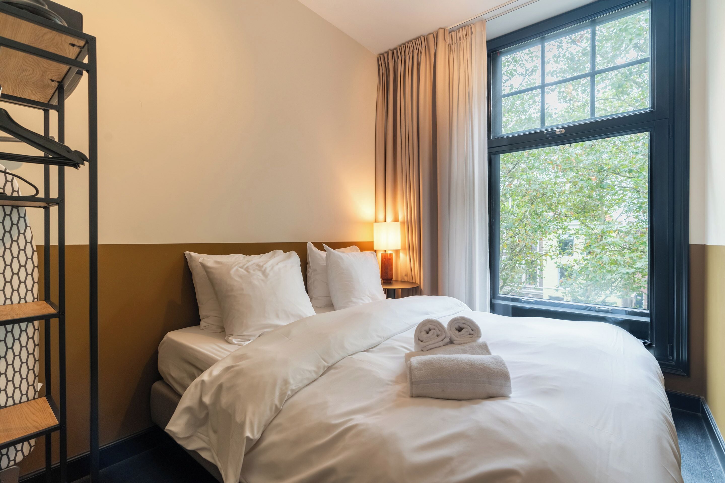 A hotel room with a bed, a bedside lamp, a window with curtains, and a shelf with a patterned item.