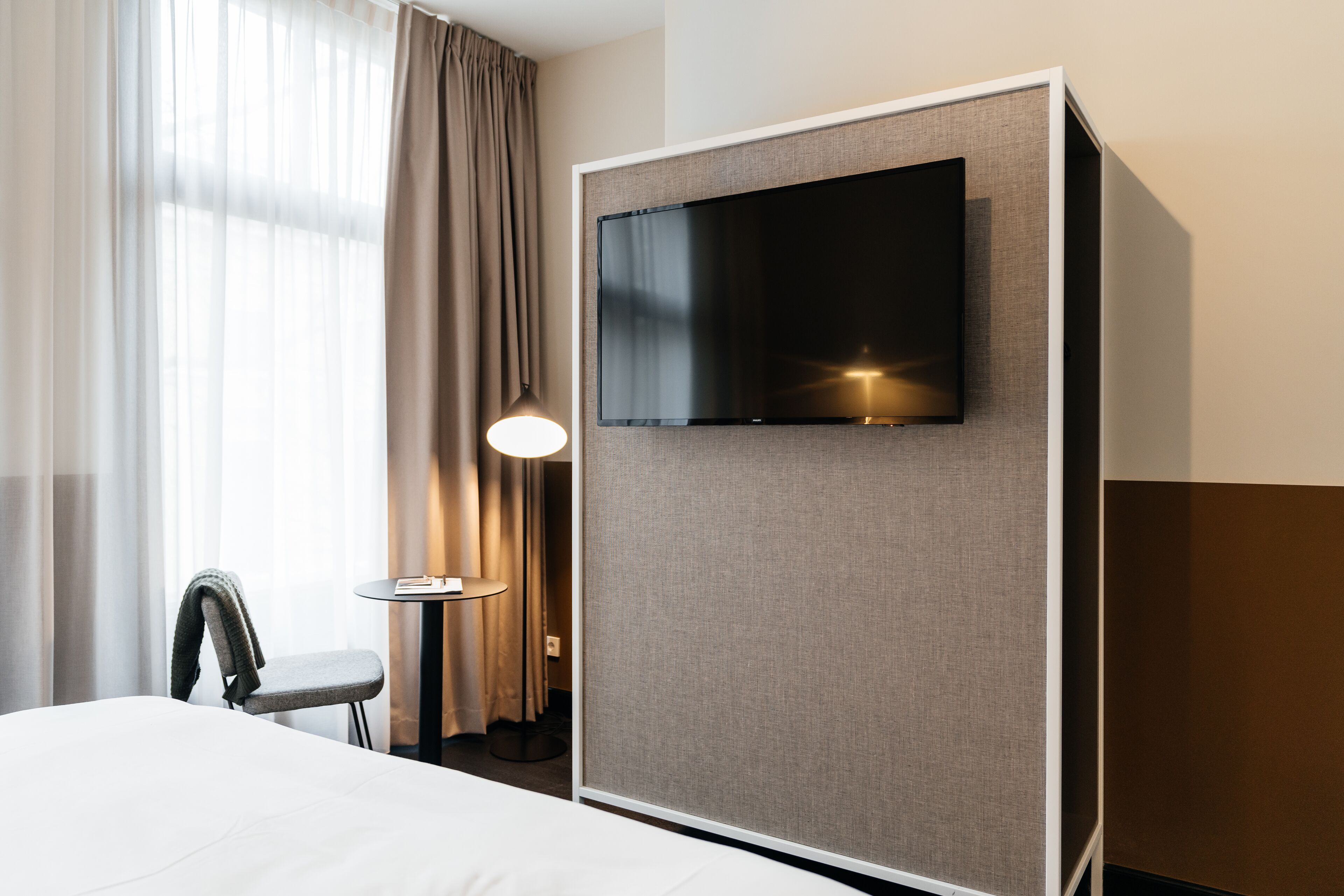 Una habitación de hotel con cama, televisor montado en la pared, una silla, una mesita con lámpara y una ventana con cortinas.