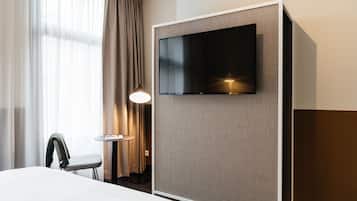 Una habitación de hotel con cama, televisor montado en la pared, una silla, una mesita con lámpara y una ventana con cortinas.