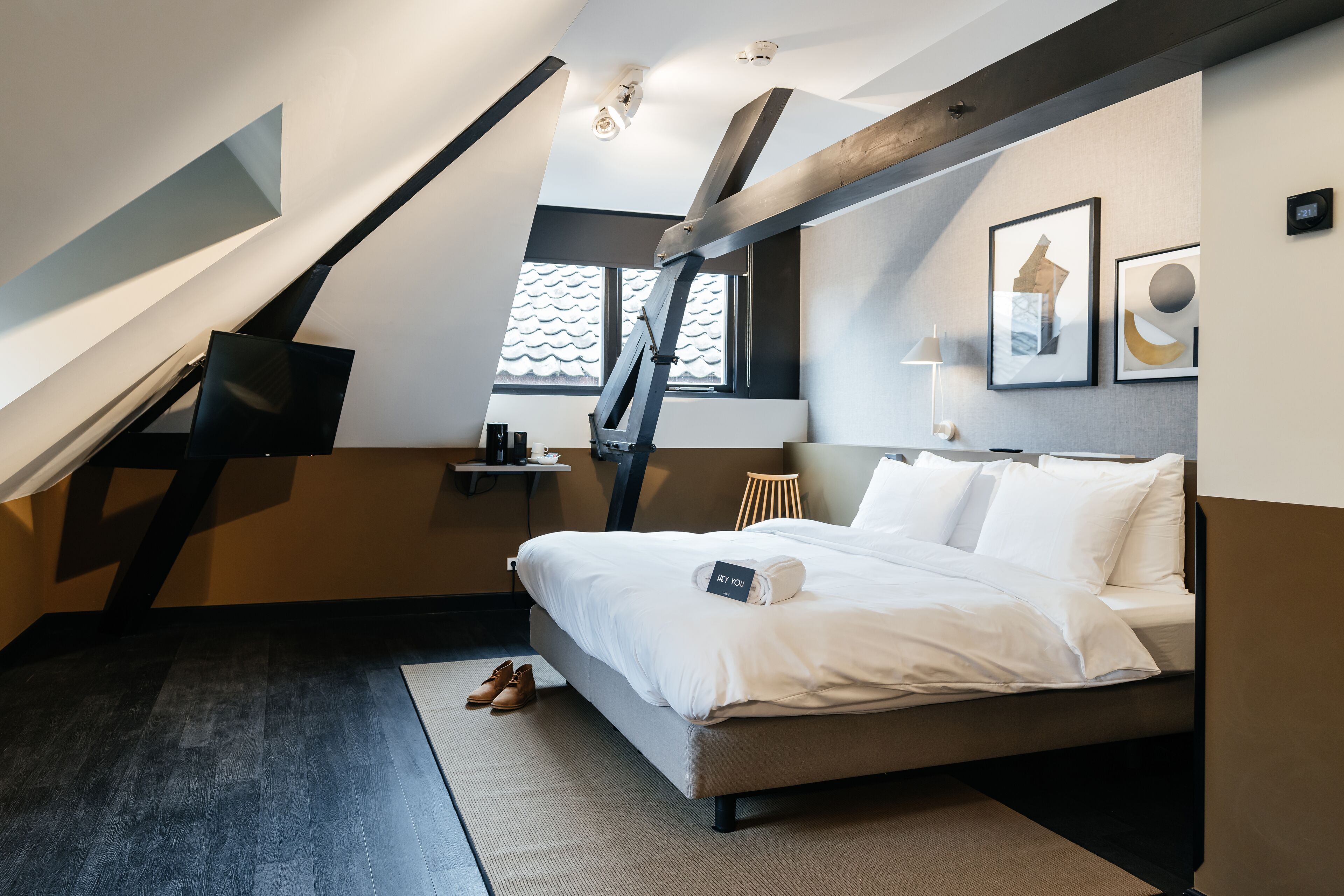 A modern hotel room with a sloped ceiling, a bed with white linens, a bedside table, a lamp, and a framed picture on the wall.