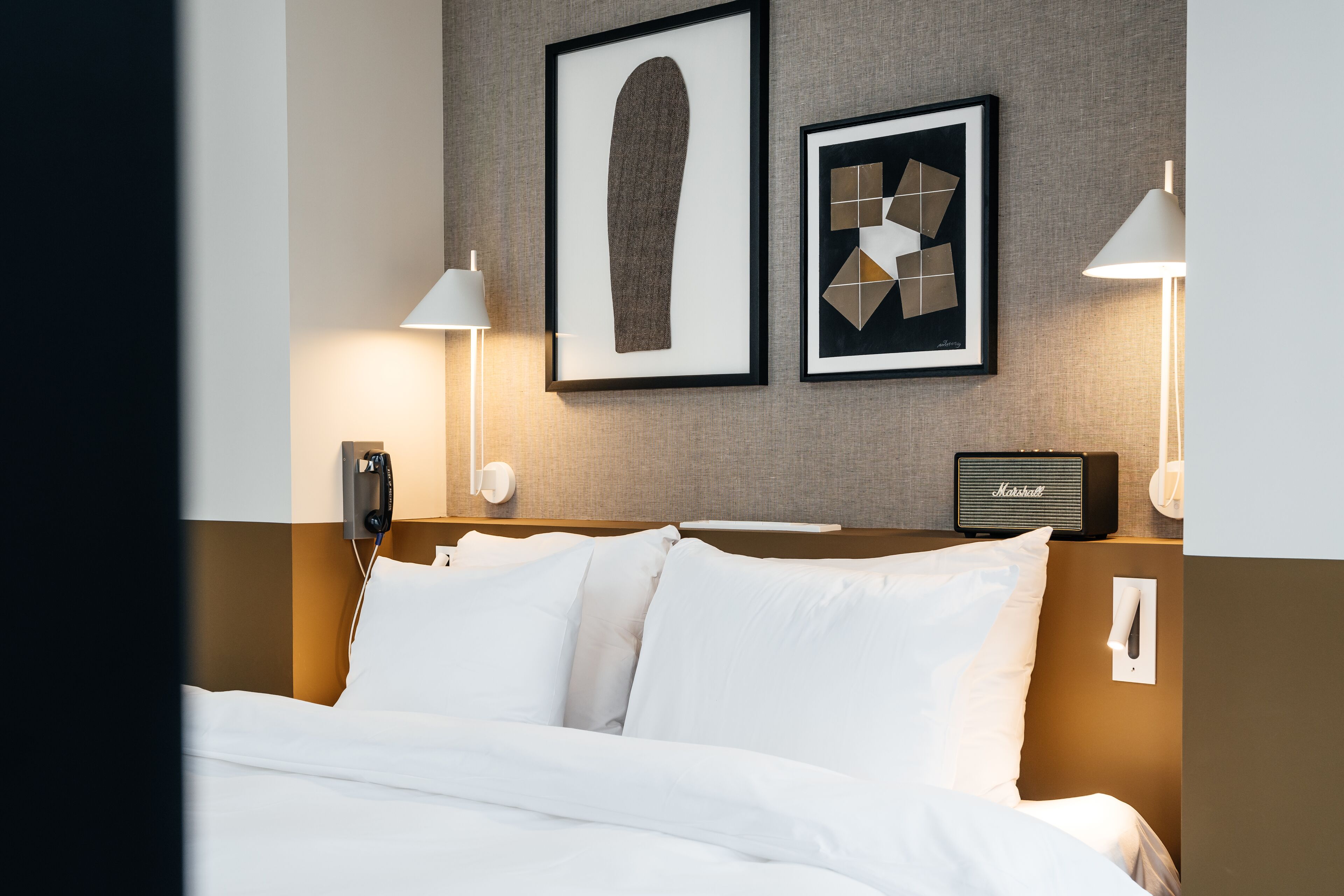 A hotel room with a neatly made bed, two framed artworks on the wall, a lamp, and a radio.