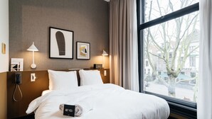 A hotel room with a large bed, two bedside lamps, a framed artwork on the wall, and a view of the city through the window. - The Hunfeld (Utrecht)