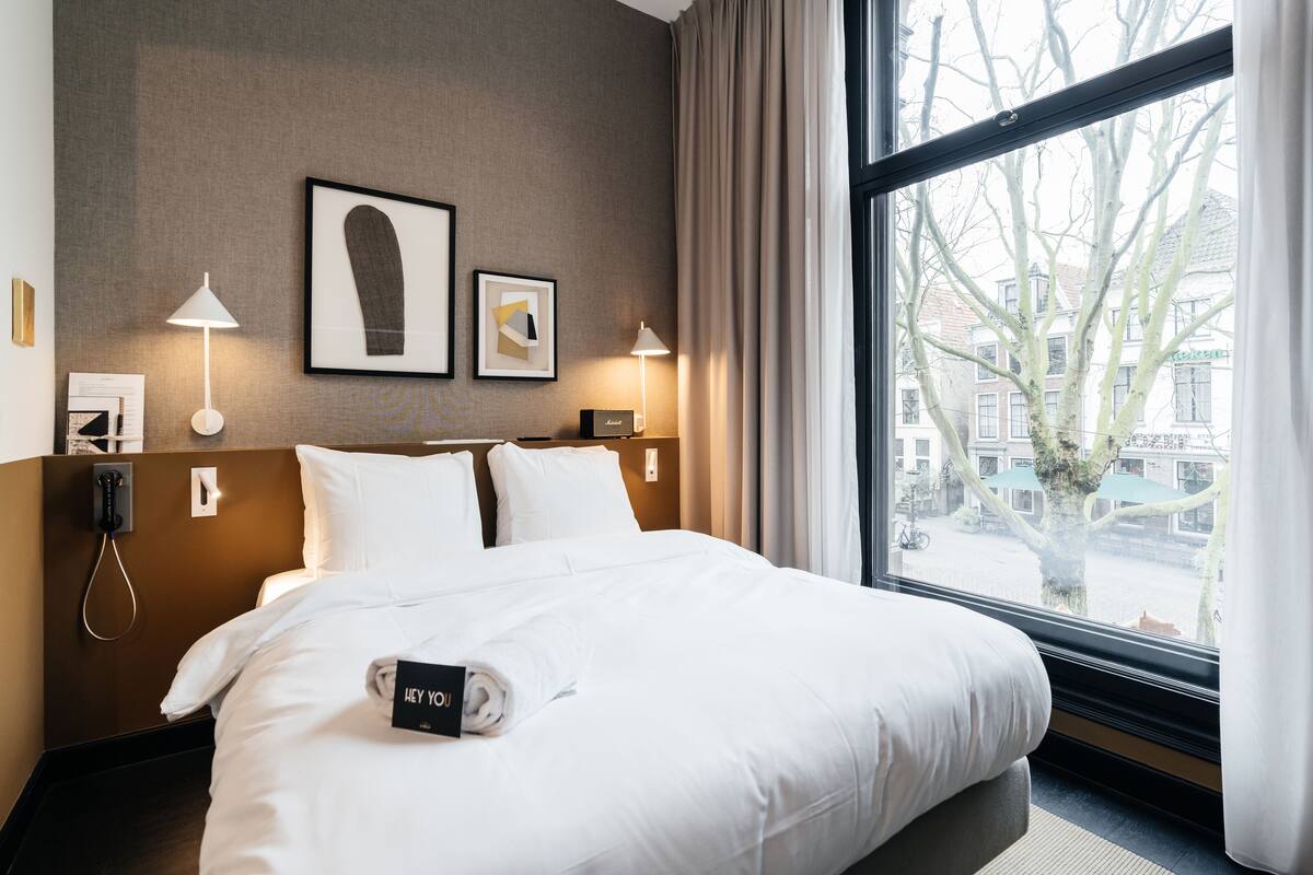 A hotel room with a large bed, two bedside lamps, a framed artwork on the wall, and a view of the city through the window.