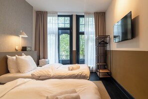 A hotel room with two beds, a TV mounted on the wall, a bedside table with a lamp, and a view of greenery through the windows.