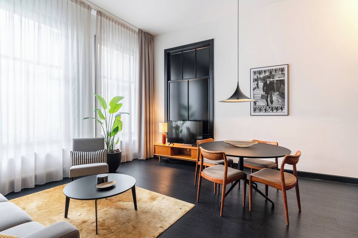 A modern living room with a sofa, a round dining table with chairs, a coffee table, a TV on a stand, a potted plant, and a framed picture on the wall.