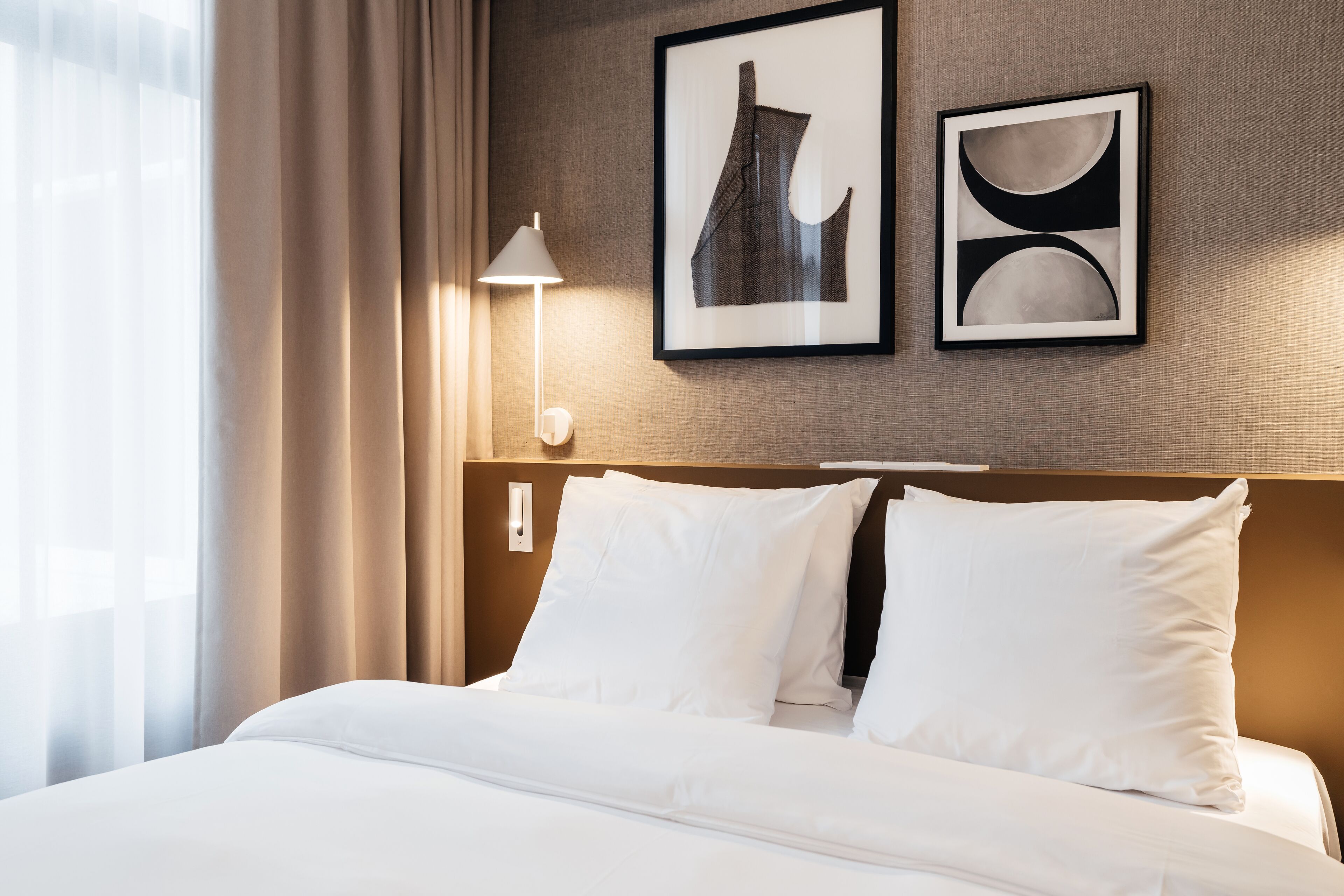 A hotel room with a neatly made bed, a lamp, and two framed artworks on the wall.