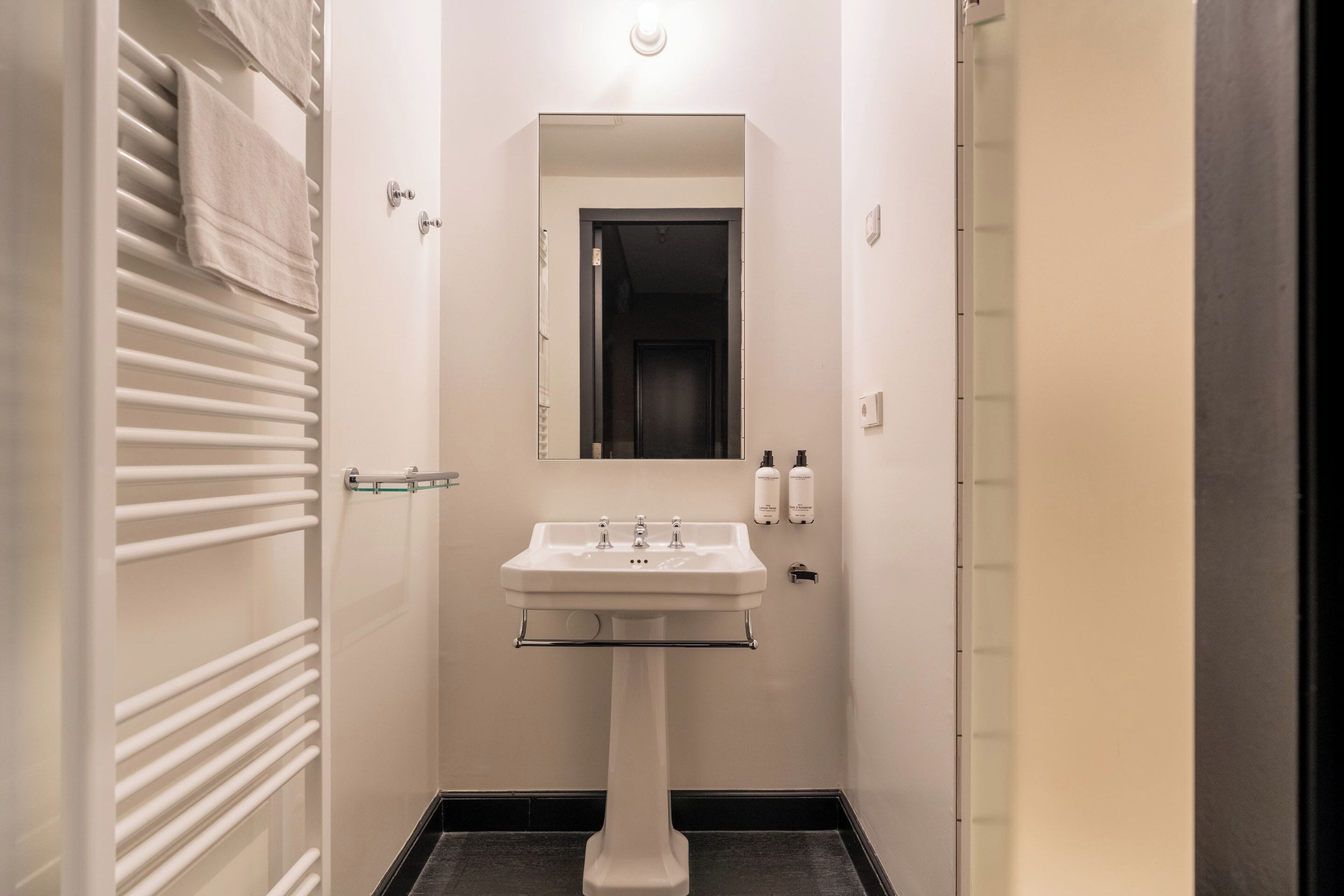 Een smalle badkamer met een wastafel op een voetstuk, een spiegel erboven en een handdoekrek aan de linkerkant.