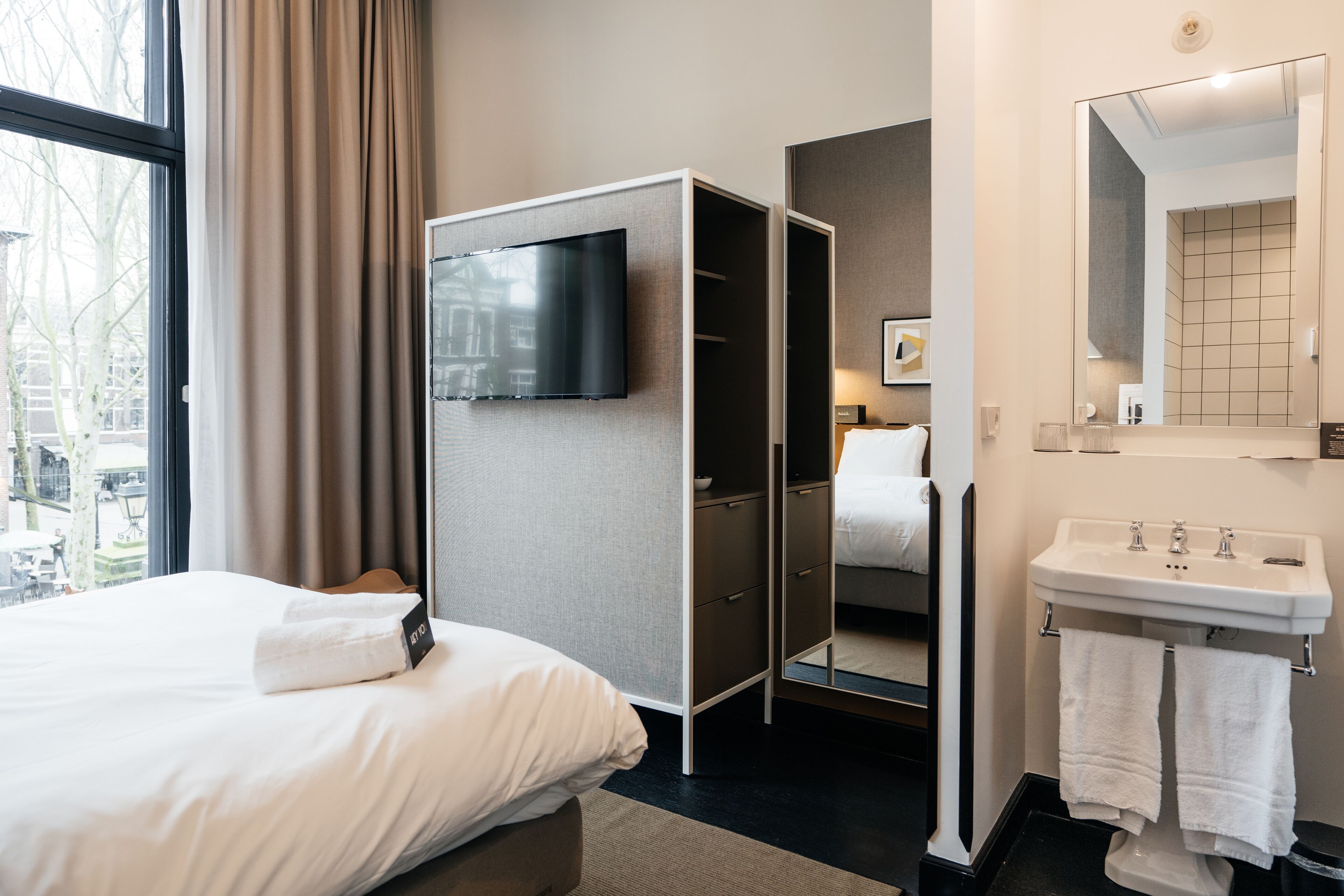 A hotel room with a bed, a wardrobe, a TV, a bathroom with a sink and mirror, and a view through a window.