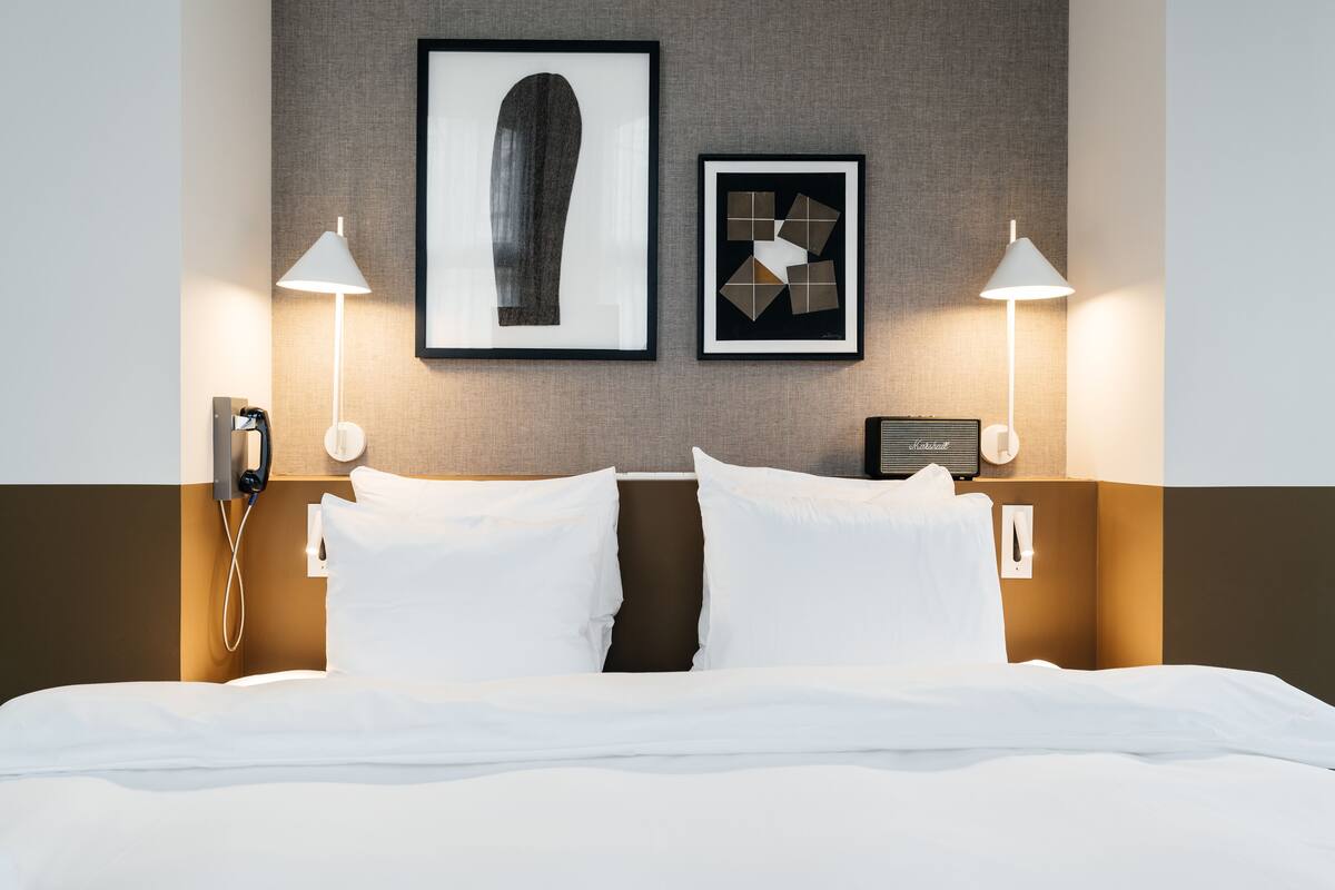 A neatly made bed with white linens, two bedside lamps, a framed artwork above the bed, and a telephone on the nightstand.