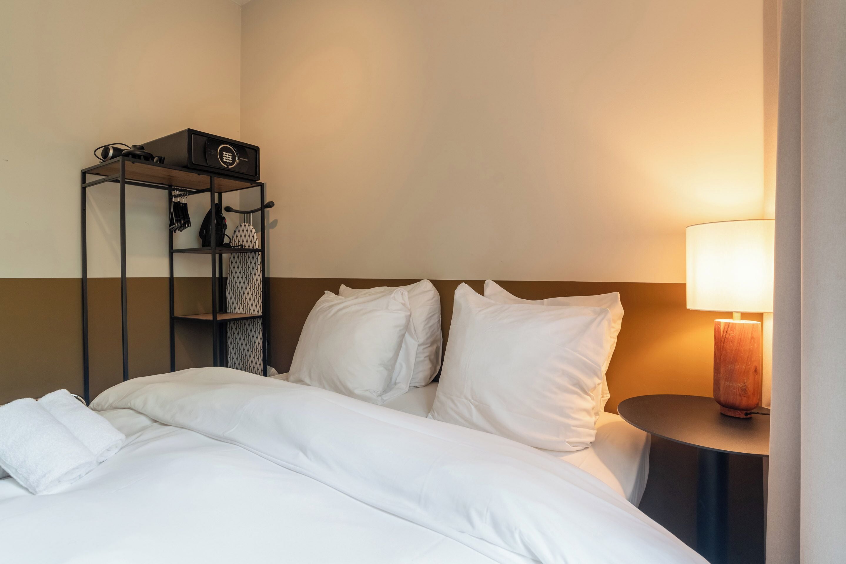 A neatly made bed with white linens, a bedside table with a lamp, and a shelf unit with a speaker and bags.