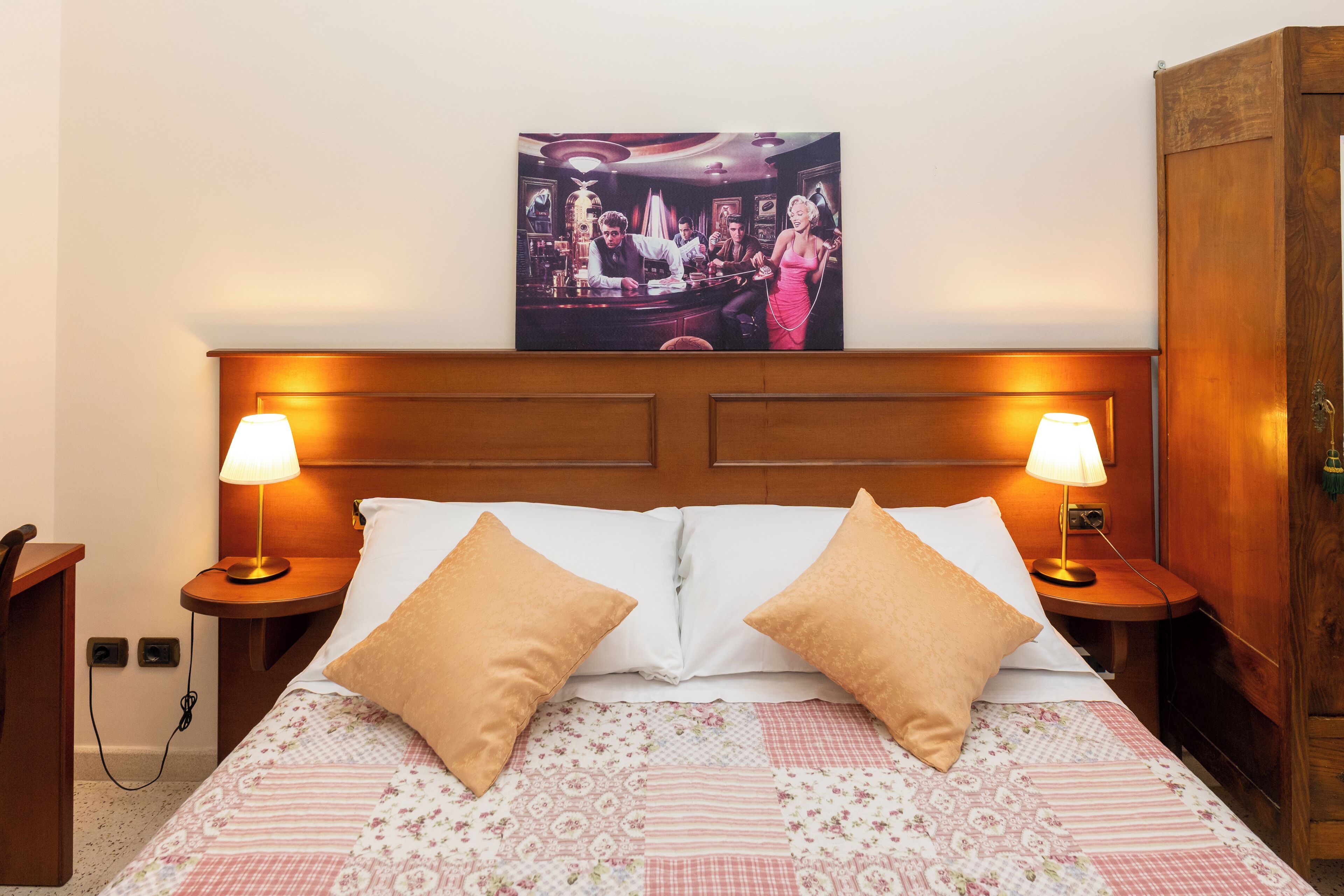 Habitación de hotel con cabecera de madera, dos lámparas de noche, cama con ropa de cama floral y un cuadro sobre la cabecera.