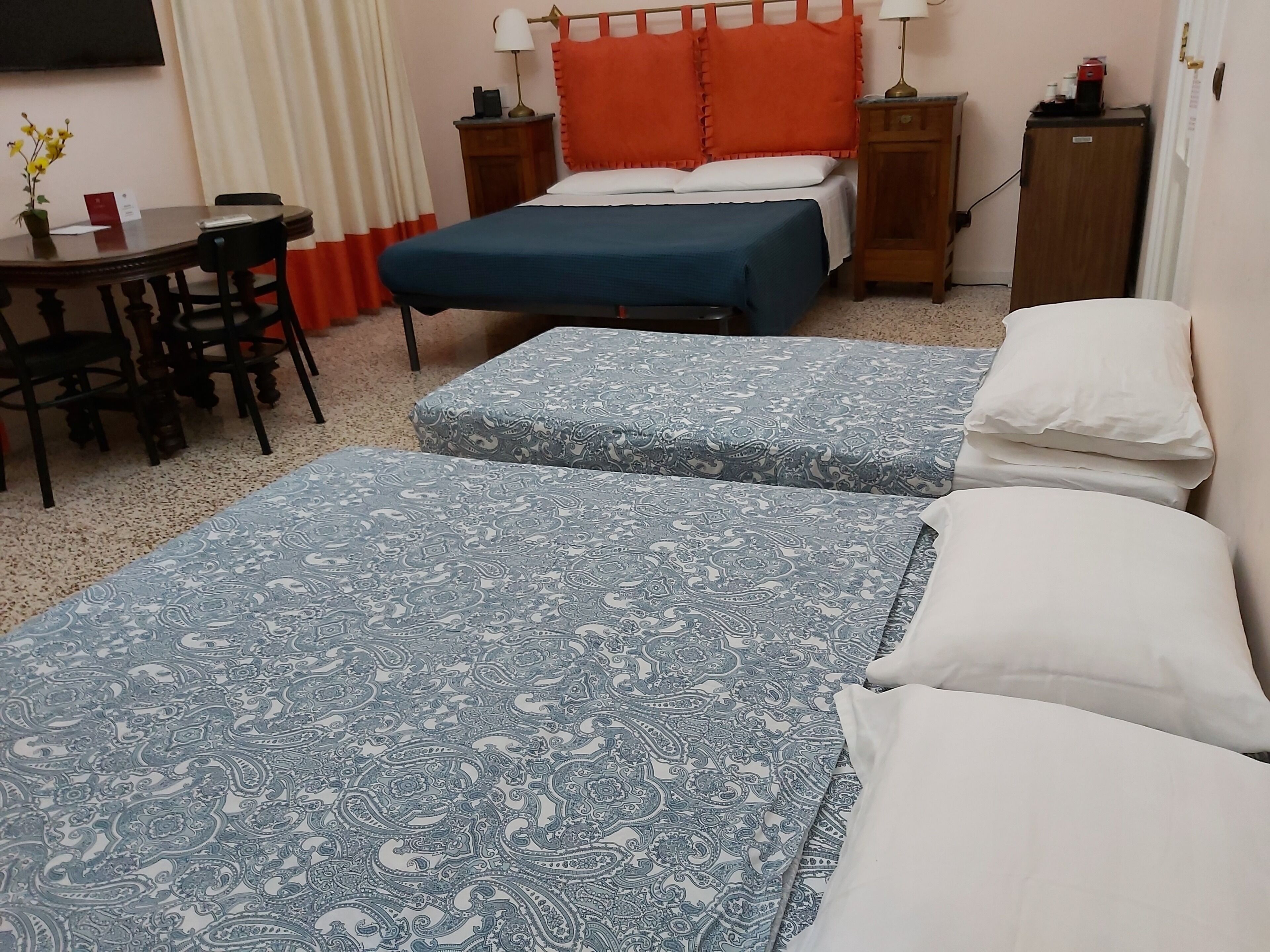 Una habitación de hotel con dos camas, una pequeña mesa con sillas, un escritorio y una mesita de noche con lámpara.