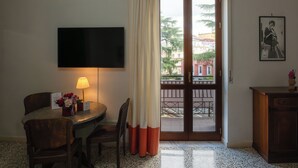 Ein Hotelzimmer mit Esstisch, Stühlen, einem flachbildfernseher an der Wand, einer Lampe, einer kleinen Blumenkomposition, einem Schrank und einer Schiebetür zum Außenbalkon mit Blick auf Gebäude und Bäume.