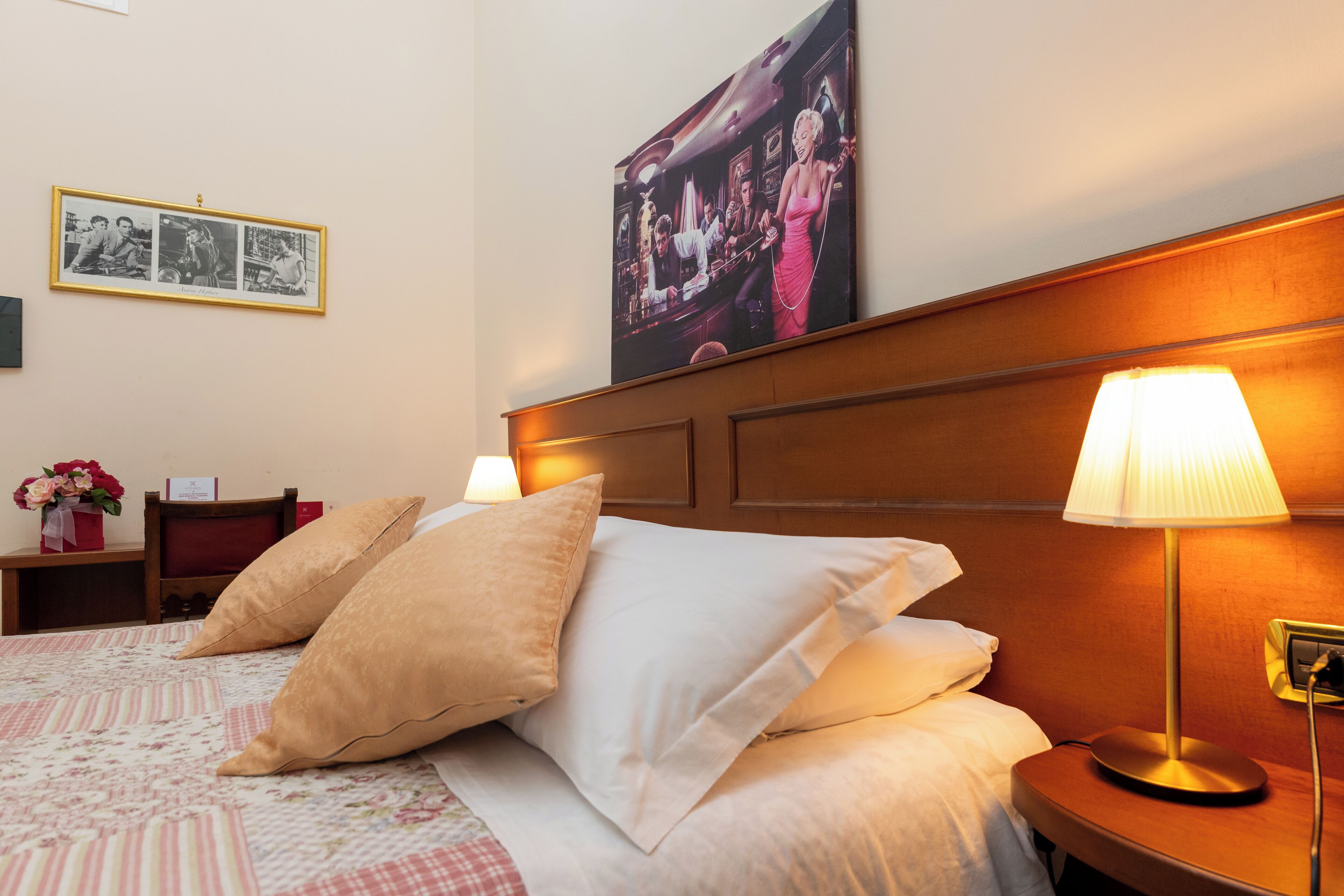 Una cama bien hecha con dos almohadas, una mesita de noche con una lámpara, un cuadro en la pared y una pequeña mesita con un florero.