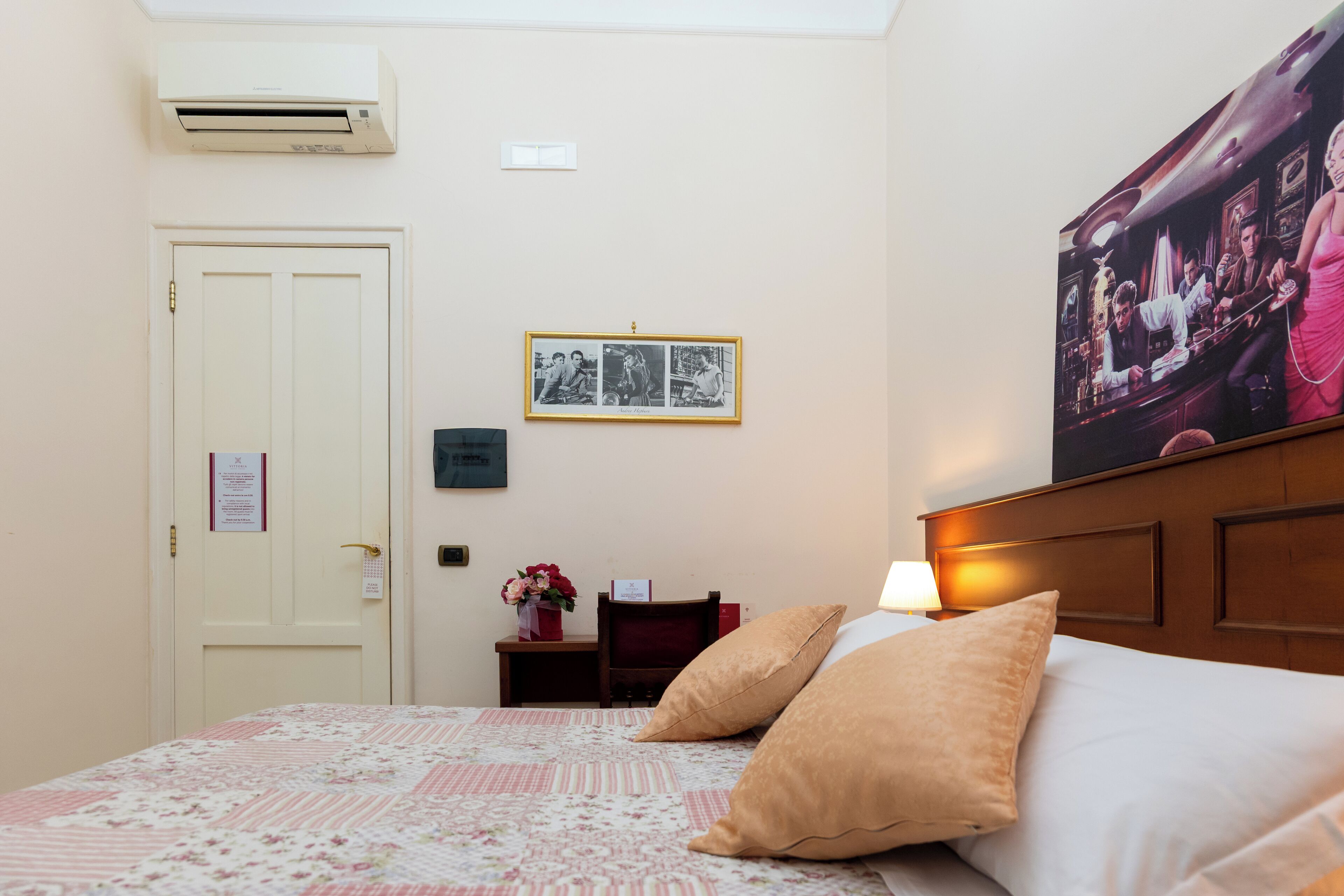 A hotel room with a bed, a nightstand, a wall-mounted air conditioner, a framed picture, and a large poster.