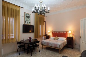 Ein Schlafzimmer mit Bett, Nachttischen, einem Fernseher, einer Chandelierleuchtre und einem Essbereich mit Tisch und Stühlen.