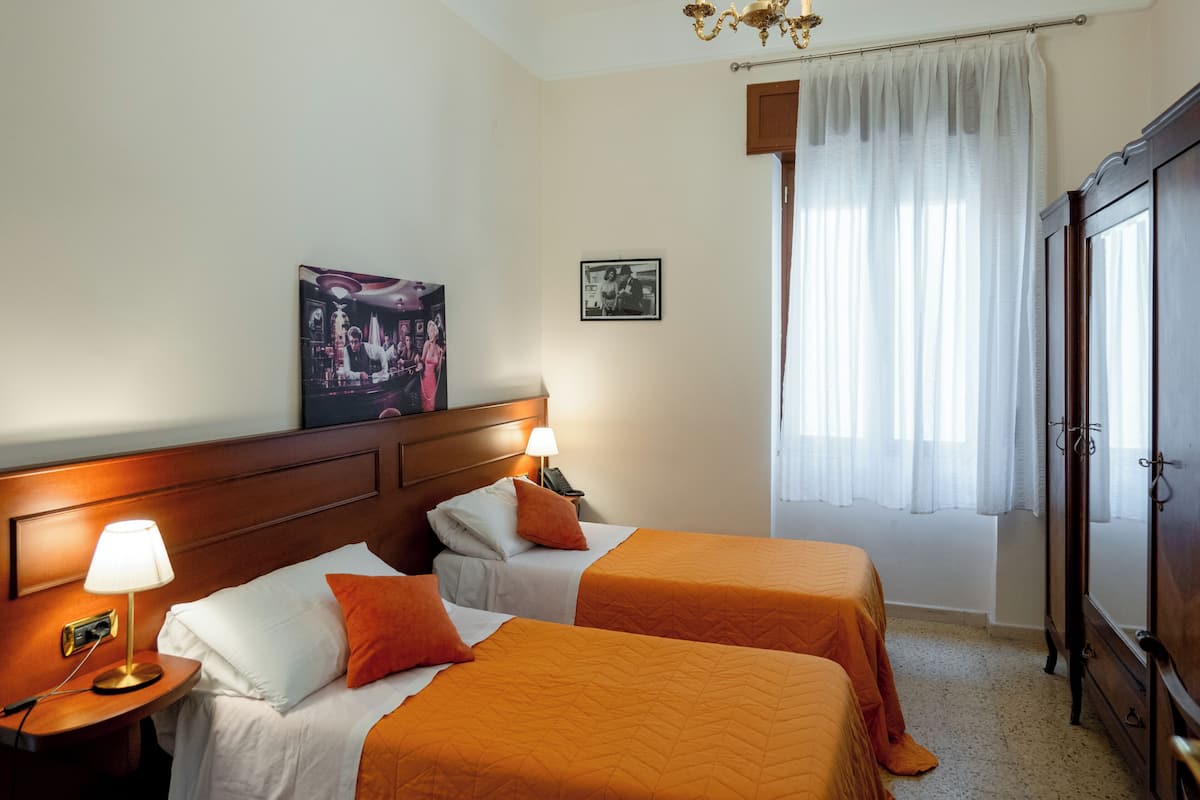Ein Hotelzimmer mit zwei Betten, einem Holzkopfteil, einem Nachttisch mit Lampe, einem Kleiderschrank und einem Fenster mit durchsichtigen Vorhängen.