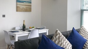 A dining area with a white table set for two, a bottle of champagne, and a bowl of fruit. There is a painting of a beach scene on the wall and a wall-mounted light fixture. - Lux Blu - Truro Apartment (Truro)